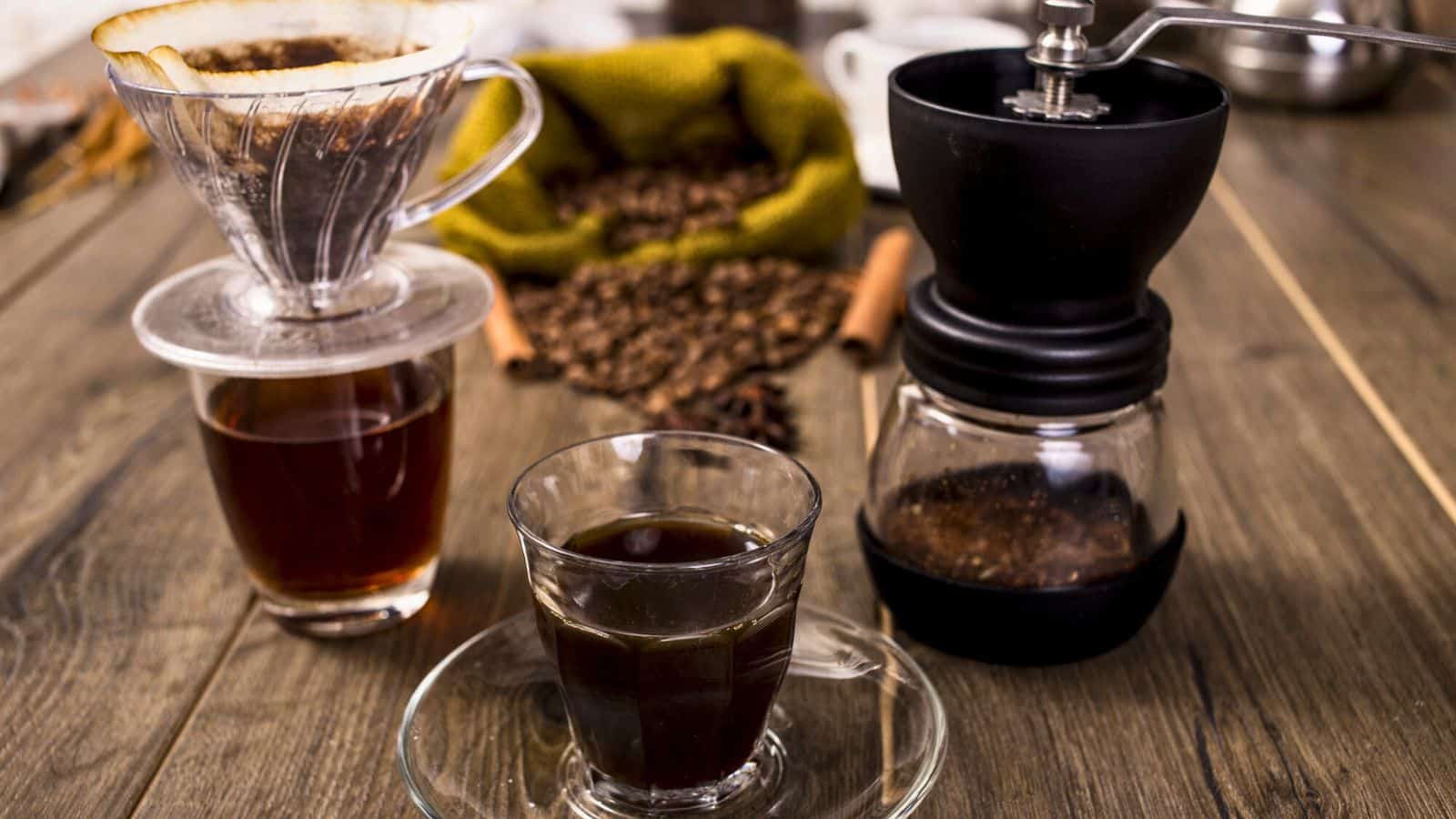 A wooden table displays a glass coffee cup and saucer, a pour-over coffee setup with a paper filter and stand, a manual coffee grinder, and a burlap sack with spilled coffee beans. Cinnamon sticks are placed nearby.
