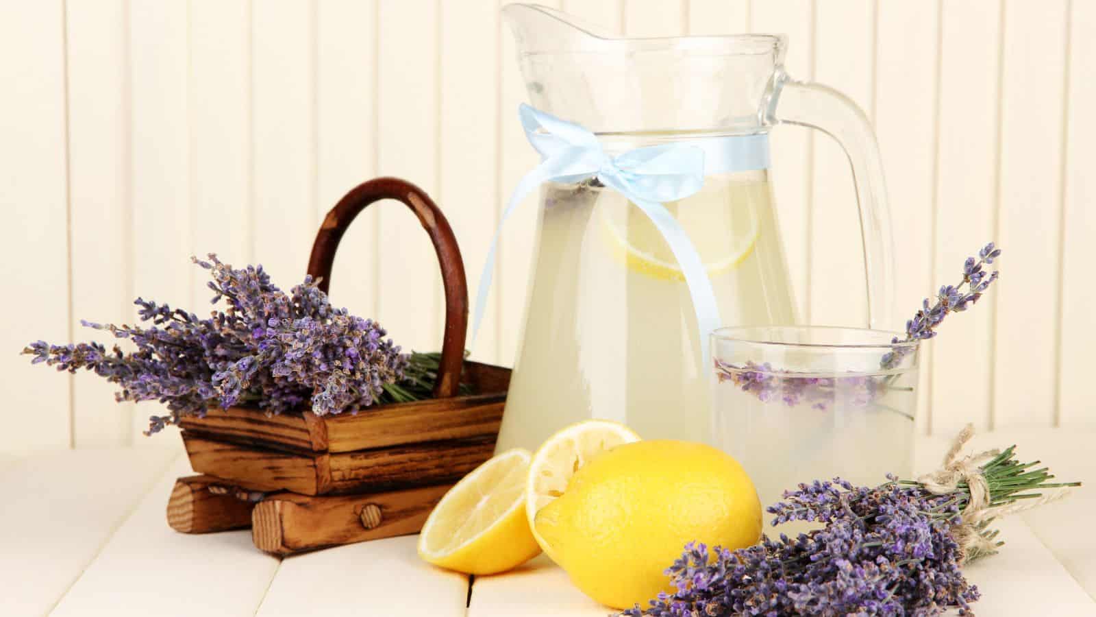 A glass pitcher filled with lemonade adorned with a blue ribbon stands on a table. A glass of lemonade is beside it. There are sprigs of lavender, a wooden basket with lavender, and halved and whole lemons on the white tabletop.
