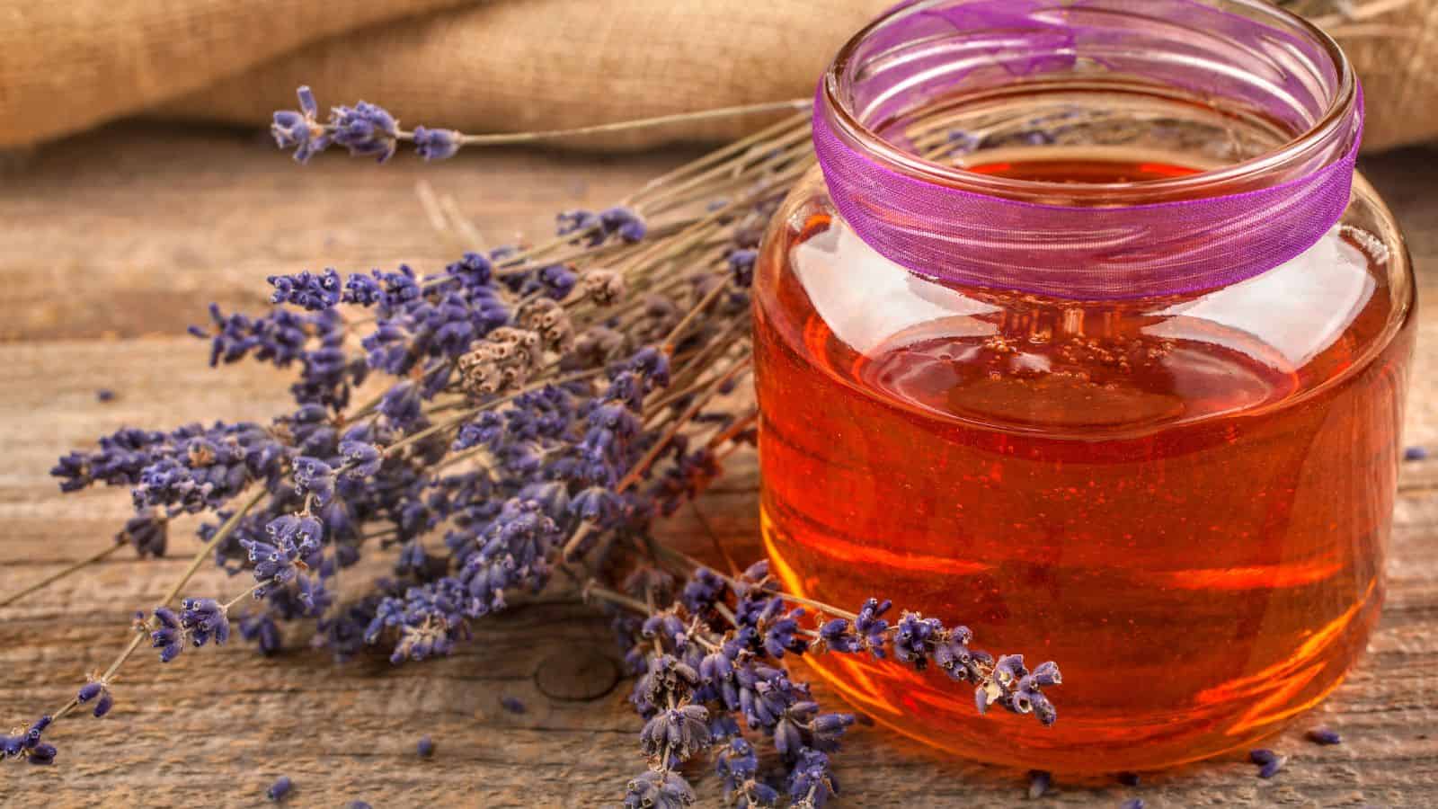 A jar filled with amber-colored liquid stands on a wooden surface. It is surrounded by sprigs of dried lavender. The jar's lid is decorated with a purple ribbon, and a burlap fabric is partially visible in the background.