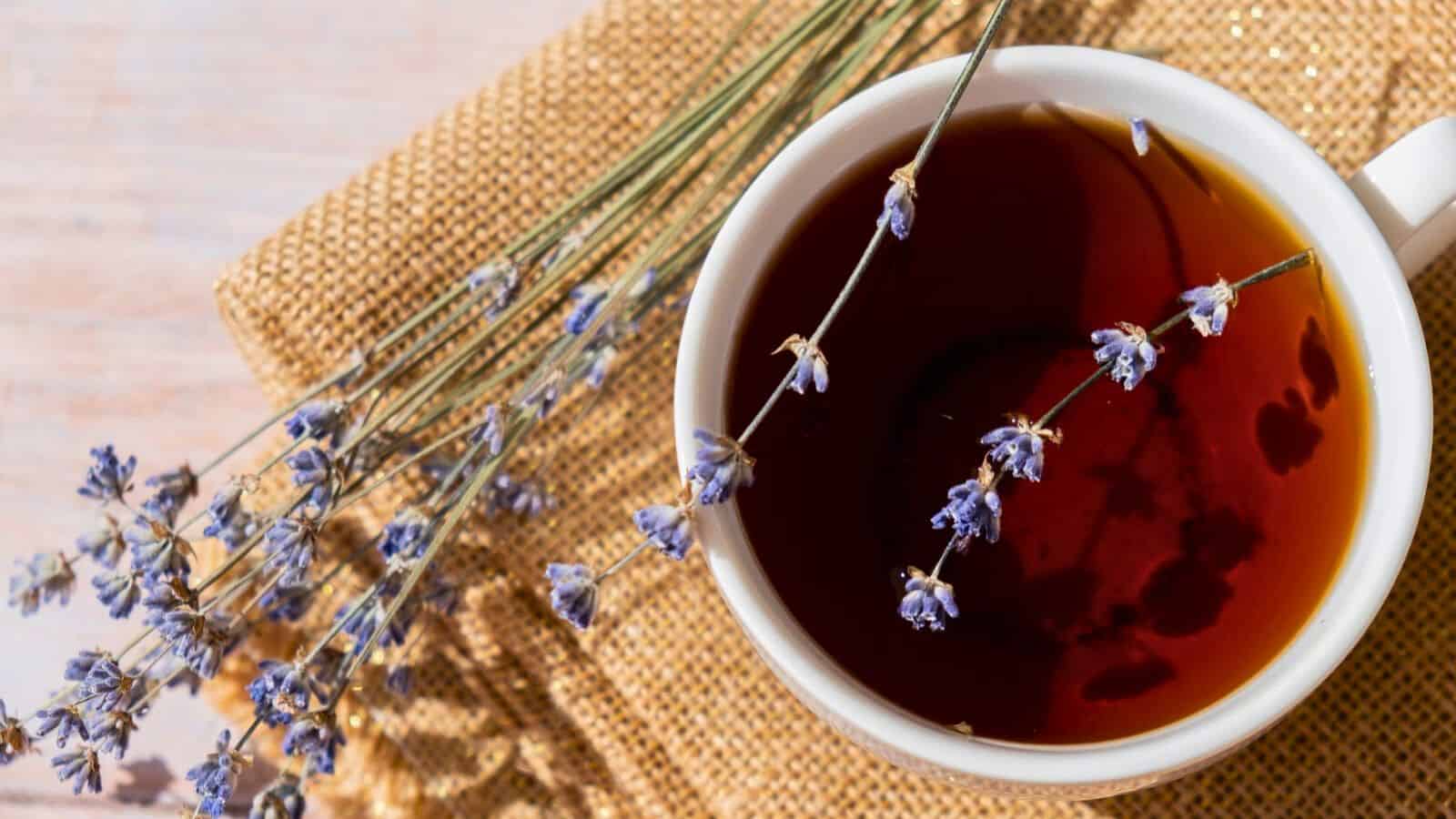 A cup of tea sits on a burlap surface, with small lavender sprigs resting across the rim. The tea is dark in color, and the lavender adds a touch of purple to the earthy tones of the setting.