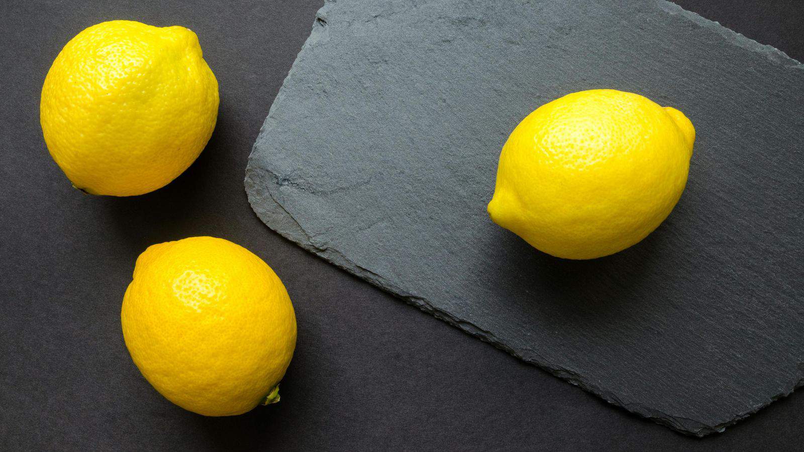 Three bright yellow lemons are arranged on a dark surface with one lemon placed on a textured slate board. The background is a rich, dark color that contrasts with the vivid lemons.