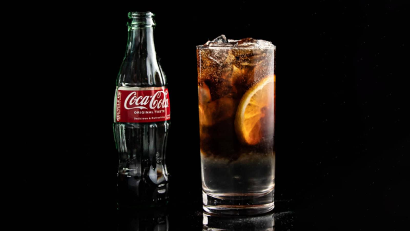 A glass filled with a dark fizzy drink and ice, garnished with an orange slice, stands on a reflective surface. A Coca-Cola bottle is placed beside the glass against a black background.