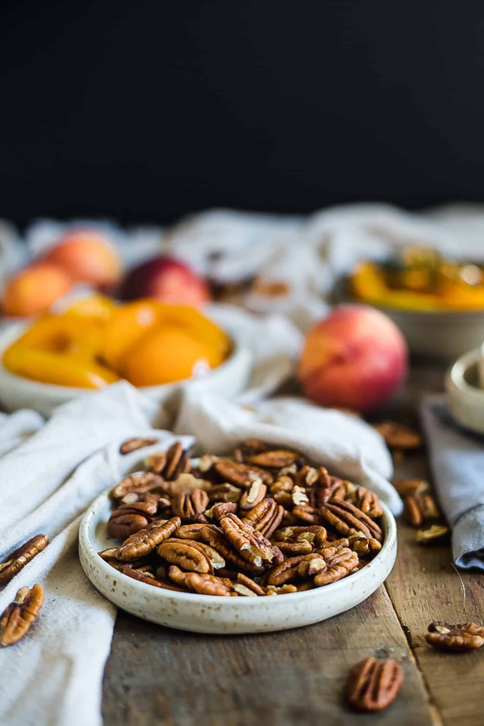 A ceramic plate filled with pecans is placed on a wooden table next to a tempting brie dip. In the background, there are whole peaches and a bowl of sliced peaches. A white cloth partially covers the table.