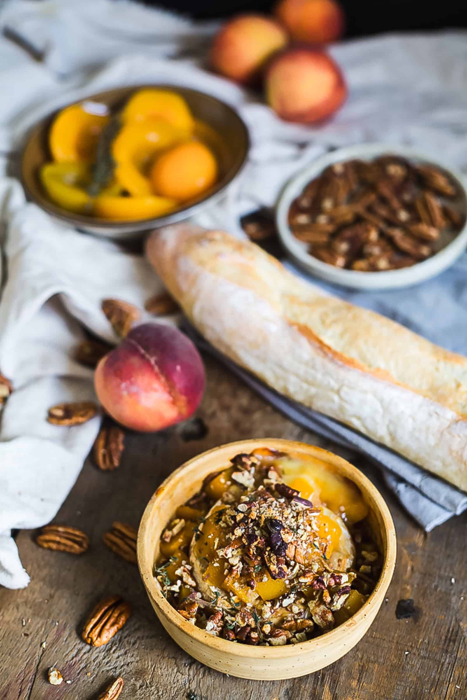 A wooden bowl of peaches topped with chopped pecans is on the table. Nearby are a baguette, a peach, and a creamy Brie dip alongside bowls of canned peaches and pecans. The items are arranged on a wooden surface with a cloth napkin.
