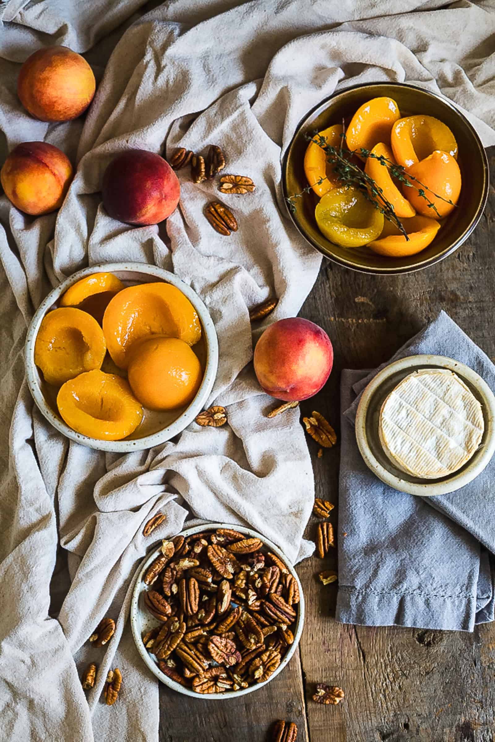 A rustic table setting features fresh peaches, canned peach halves in bowls, pecans, and a round of soft cheese on a cloth. White and blue linens are draped around the items, highlighting a creamy Brie dip ready to complement the spread.