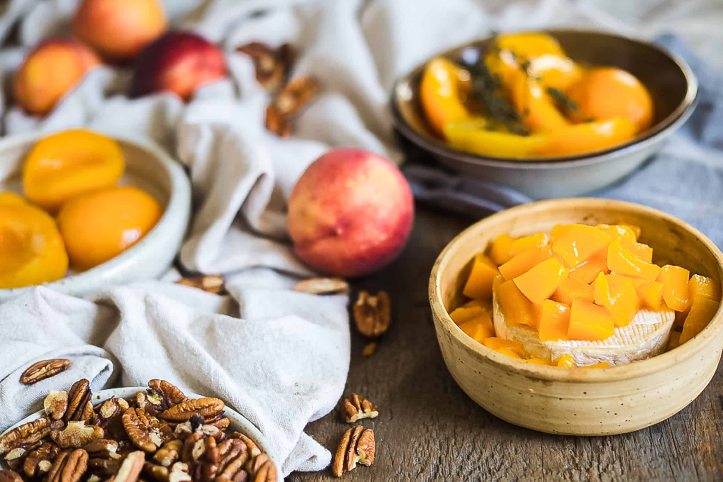 Bowls containing halved peaches, sliced peaches, and chopped peaches with brie are on a wooden surface. Whole peaches, pecans, and a fabric cloth are scattered around the bowls, creating a perfect setup for a delightful brie dip experience.