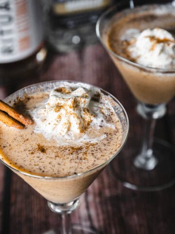 Two creamy cocktails with frothy tops are garnished with cinnamon sticks and sprinkled spice in cocktail glasses on a wooden surface. A bottle in the background and a small bowl with frothy cream are partially visible.