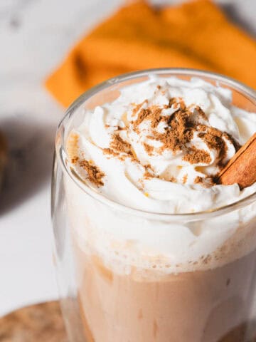 A glass mug filled with a whipped cream-topped beverage, garnished with a sprinkle of cinnamon and a cinnamon stick. A small pumpkin and an orange cloth are in the blurred background on a marble surface.