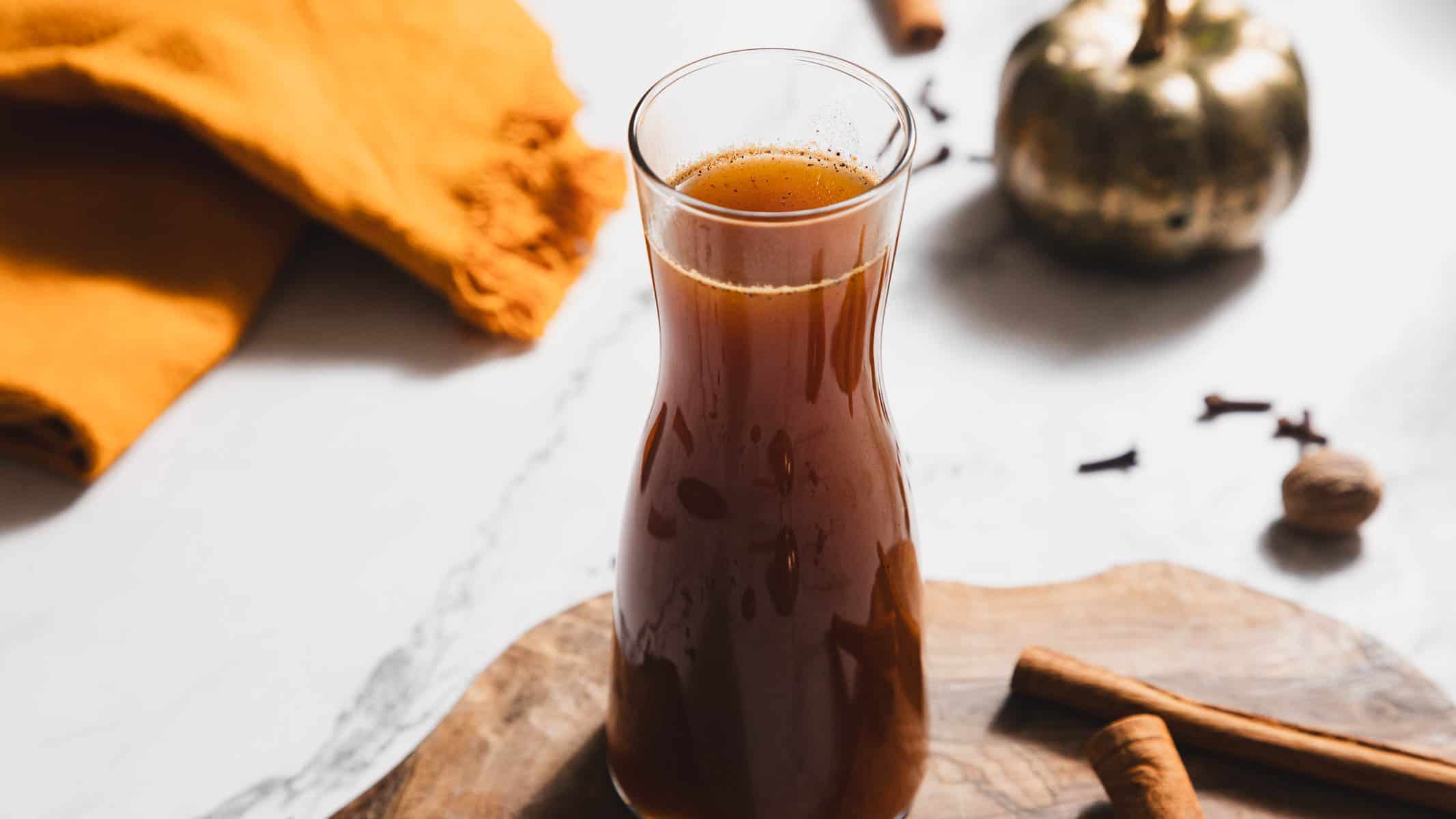 A tall glass carafe filled with dark apple cider sits on a wooden board, accompanied by cinnamon sticks. Nearby, a brass-colored decorative pumpkin and an orange cloth create cozy vibes, while cloves and nutmeg add spice. A hint of pumpkin spice syrup aroma completes the autumn scene.