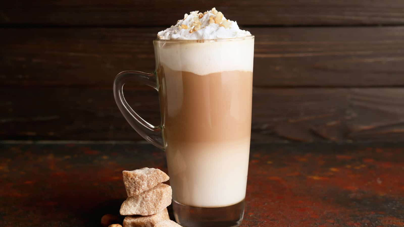 A tall glass mug filled with a layered coffee drink, topped with whipped cream and sprinkled with crushed nuts. Three pieces of brown sugar sit beside the mug on a dark, textured surface with a wooden background.