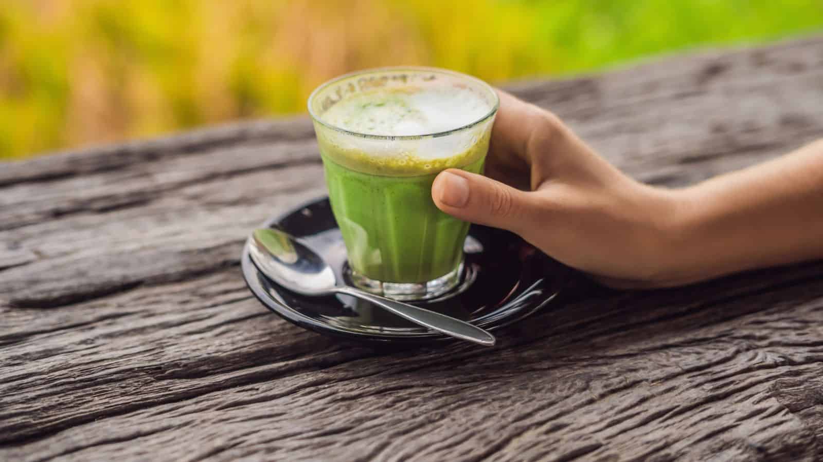 A hand holds a small glass of green matcha latte, placed on a black saucer with a spoon, resting on a rustic wooden surface. The background is blurred with greenery.