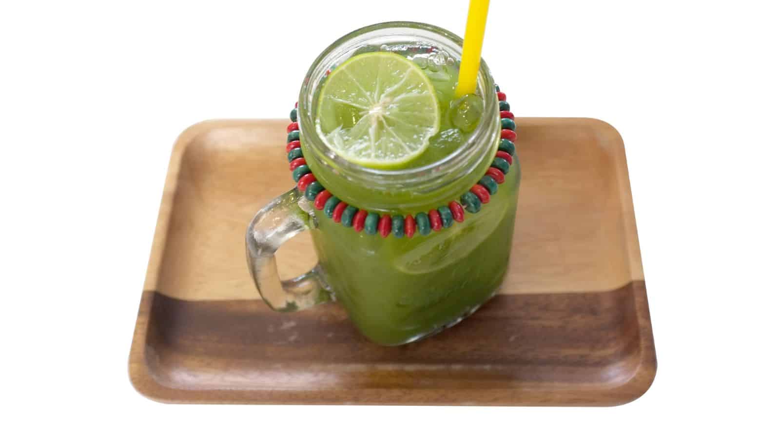 A glass mug filled with a green drink, topped with two lime slices. The mug has a yellow straw and a beaded decoration around the rim. It is placed on a wooden tray.