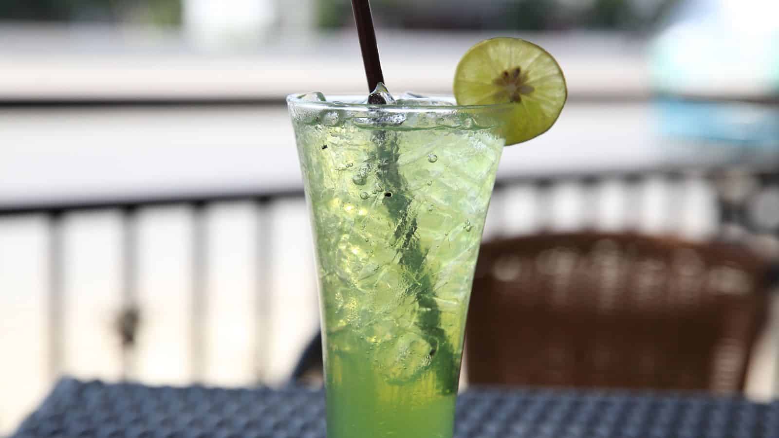 A tall glass of green iced drink with ice cubes and a black straw, garnished with a slice of lime on the rim. The glass is on a textured table with blurred outdoor seating in the background.