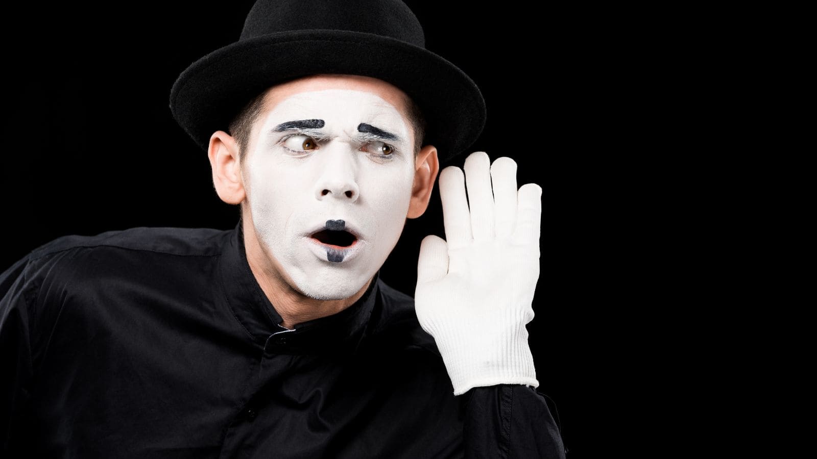 A mime artist in a black hat and outfit gestures with a hand to his ear. His face is painted white with black accents, and he wears white gloves. The background is black, enhancing his theatrical expression.