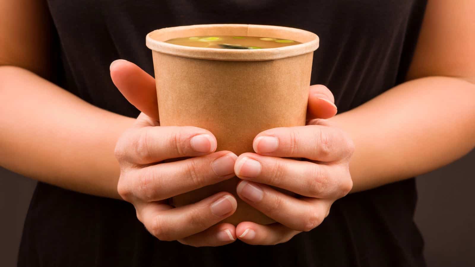 A person holds a brown paper cup filled with soup, garnished with green herbs, against a dark background. The person's hands are visible, but their face and surroundings are not shown.
