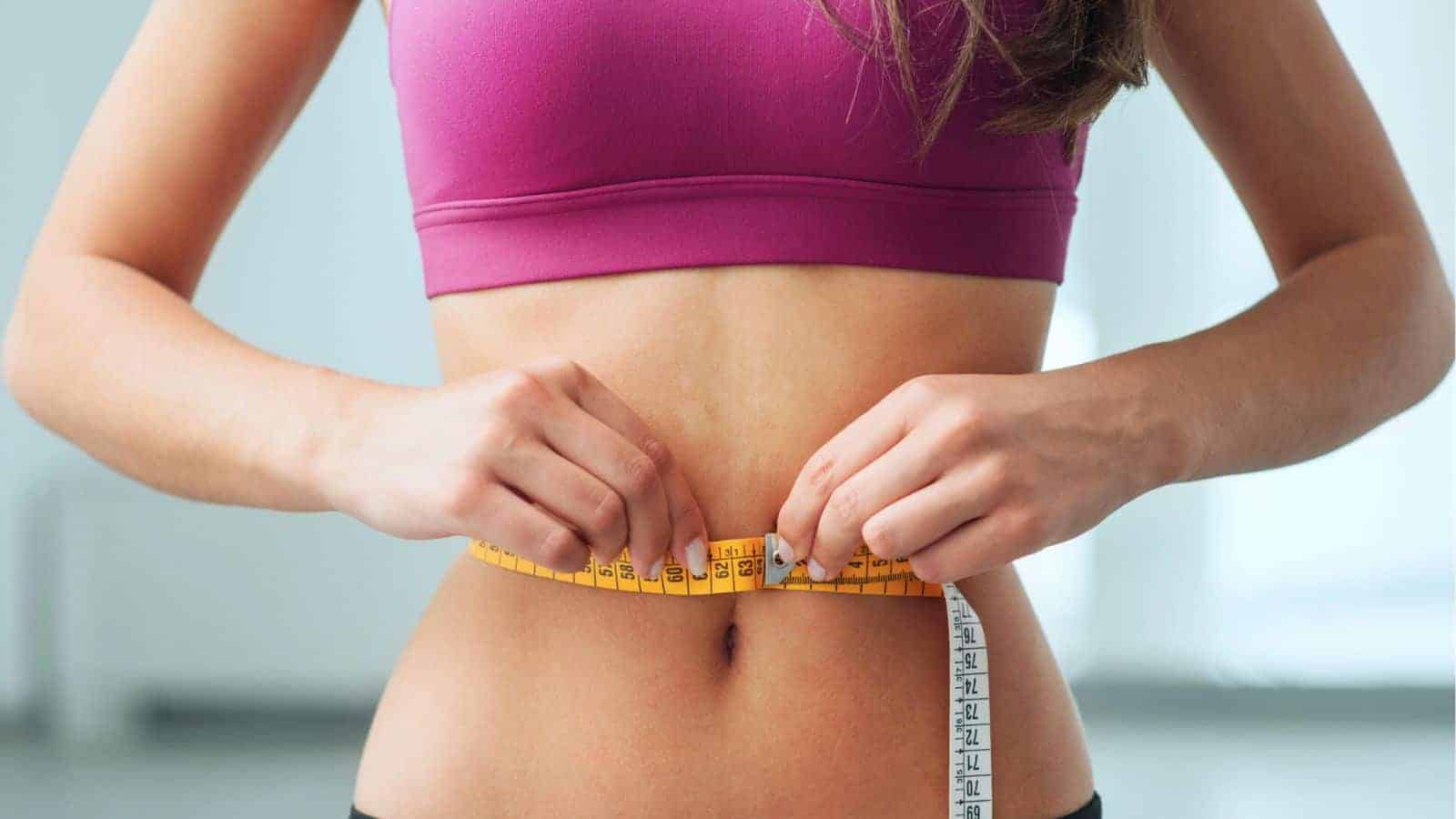 A person in a pink sports top is measuring their waist with a yellow tape measure. The background is softly blurred.
