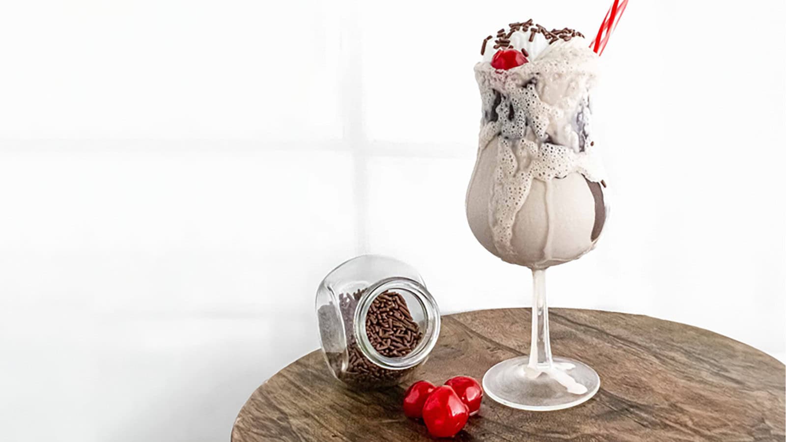 A creamy milkshake topped with chocolate sprinkles and a cherry in a tall glass with a red straw. Next to it is an overturned jar of chocolate sprinkles and two cherries on a wooden table against a white background.