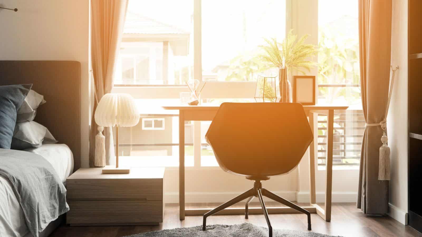 A sunlit bedroom features a neatly made bed with gray bedding, a wooden desk with a black chair facing a large window, and a potted plant on the desk. A soft light filters through the sheer curtains, creating a warm and inviting atmosphere.