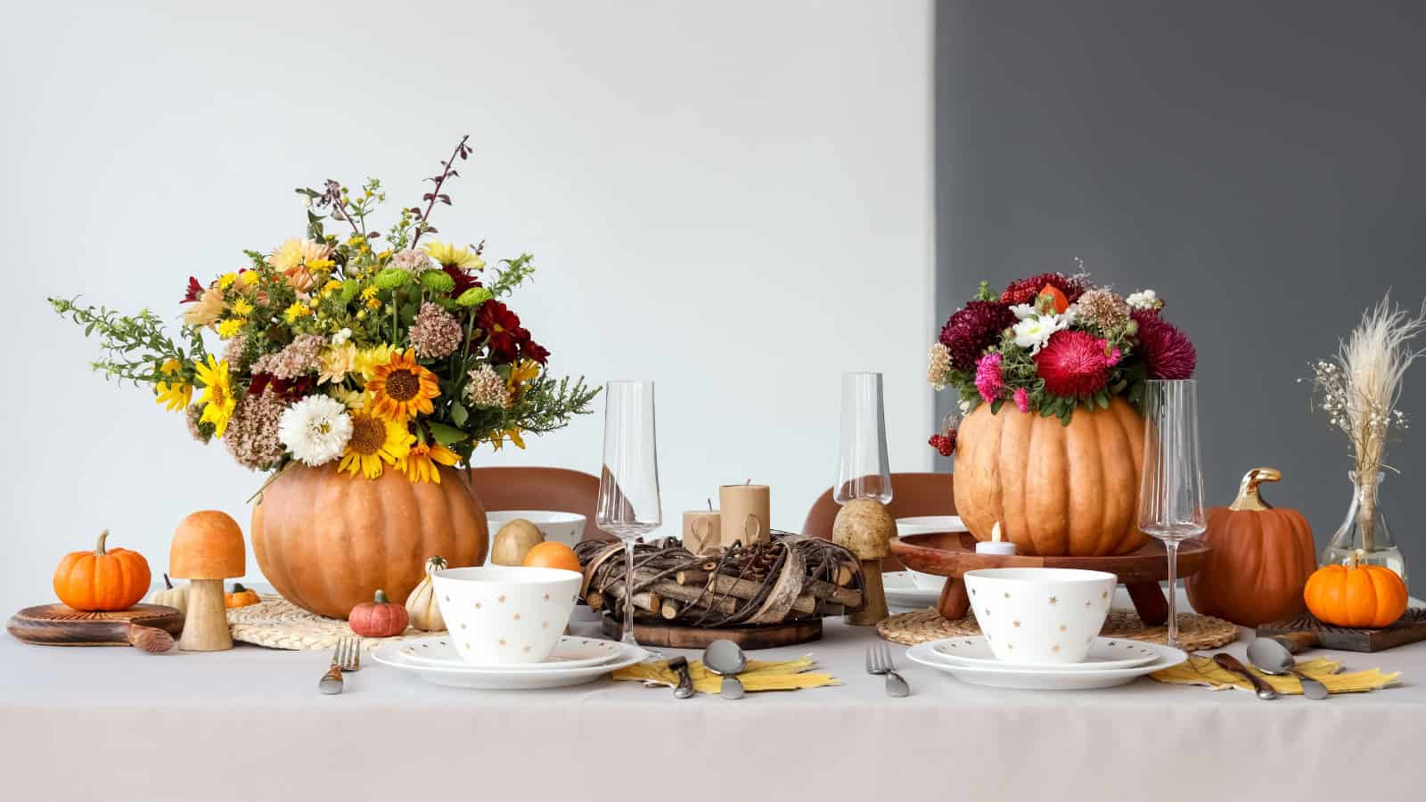 A dining table set for fall, featuring two pumpkin vases with vibrant floral arrangements, decorative wooden items, small pumpkins, and elegant tableware with polka dots. The setting includes candles and a mix of seasonal decor.