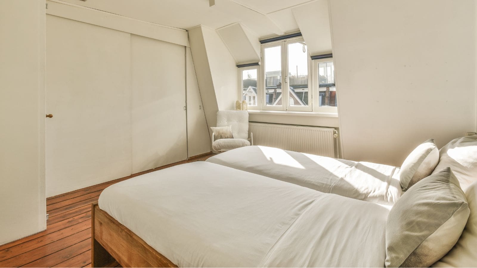 A minimalist bedroom featuring two twin beds with white bedding, a light grey armchair by the window, and wooden flooring. The room has a white closet with sliding doors and a large window allowing natural light to enter.