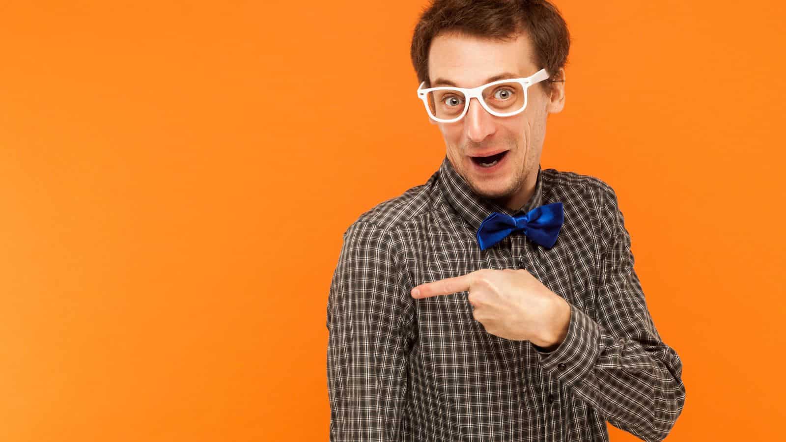 A man with white glasses and a blue bow tie, wearing a plaid shirt, points to his right side with an excited expression. The background is solid orange.