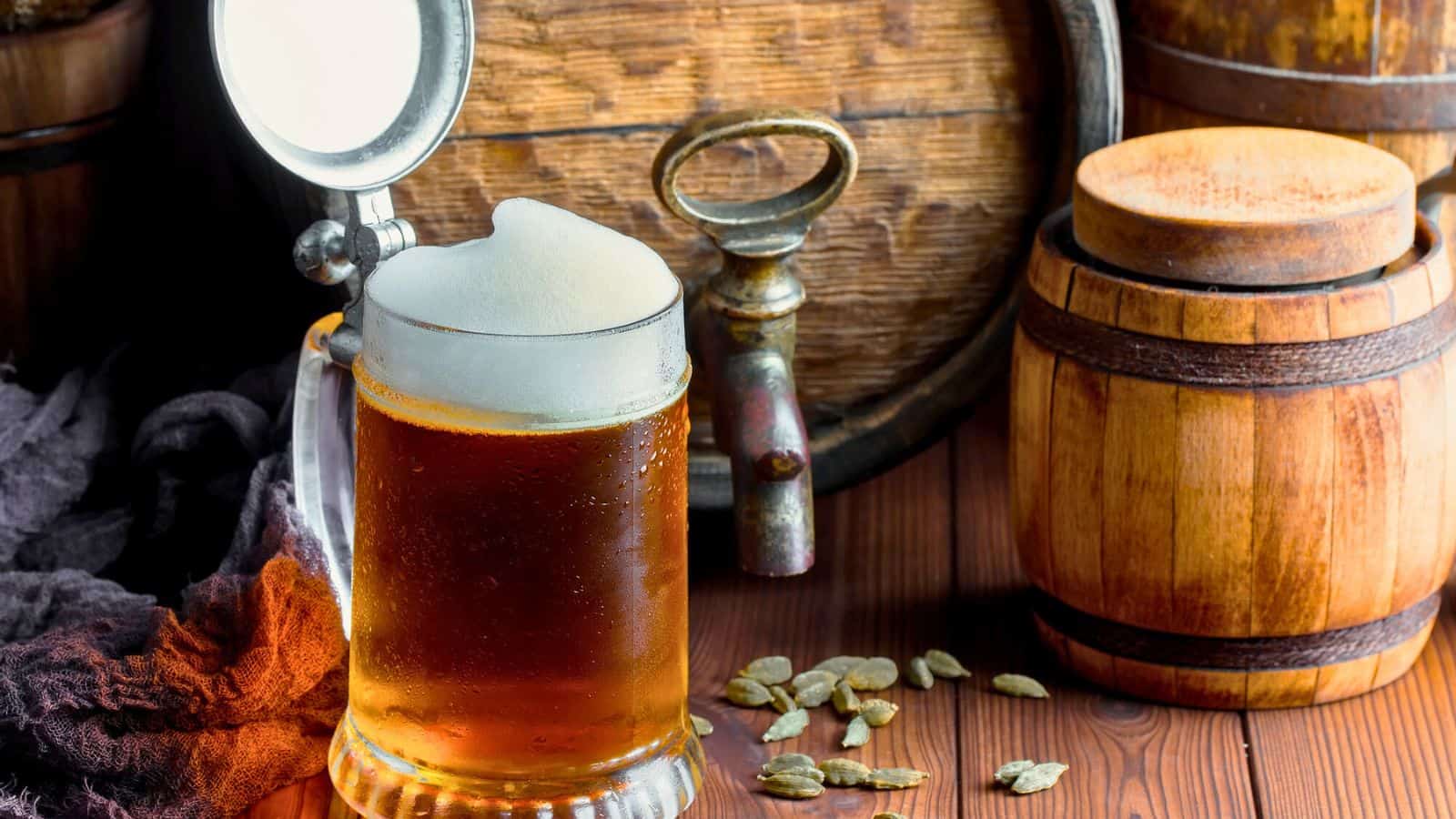 A frothy glass of beer sits on a wooden surface next to a small wooden keg. Barley grains are scattered nearby. A vintage beer tap is visible in the background.
