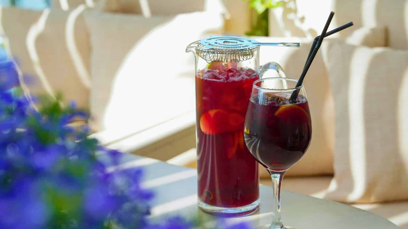 A glass pitcher and a wine glass are filled with red sangria, garnished with slices of fruit. Both are placed on a table with a blurred background of blue flowers and cushions. Sunlight streams through, creating a bright and inviting atmosphere.