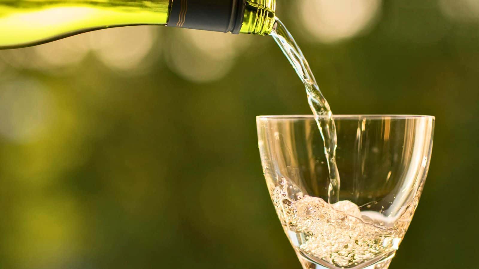 A clear liquid is being poured from a green wine bottle into a transparent wine glass, set against a blurred green background.
