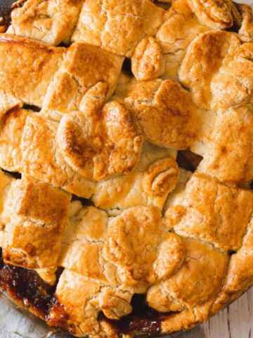 A freshly baked pie with a golden, lattice crust sits on a wooden surface. A gray cloth is draped nearby. A few slices of green apple are visible in the corner.