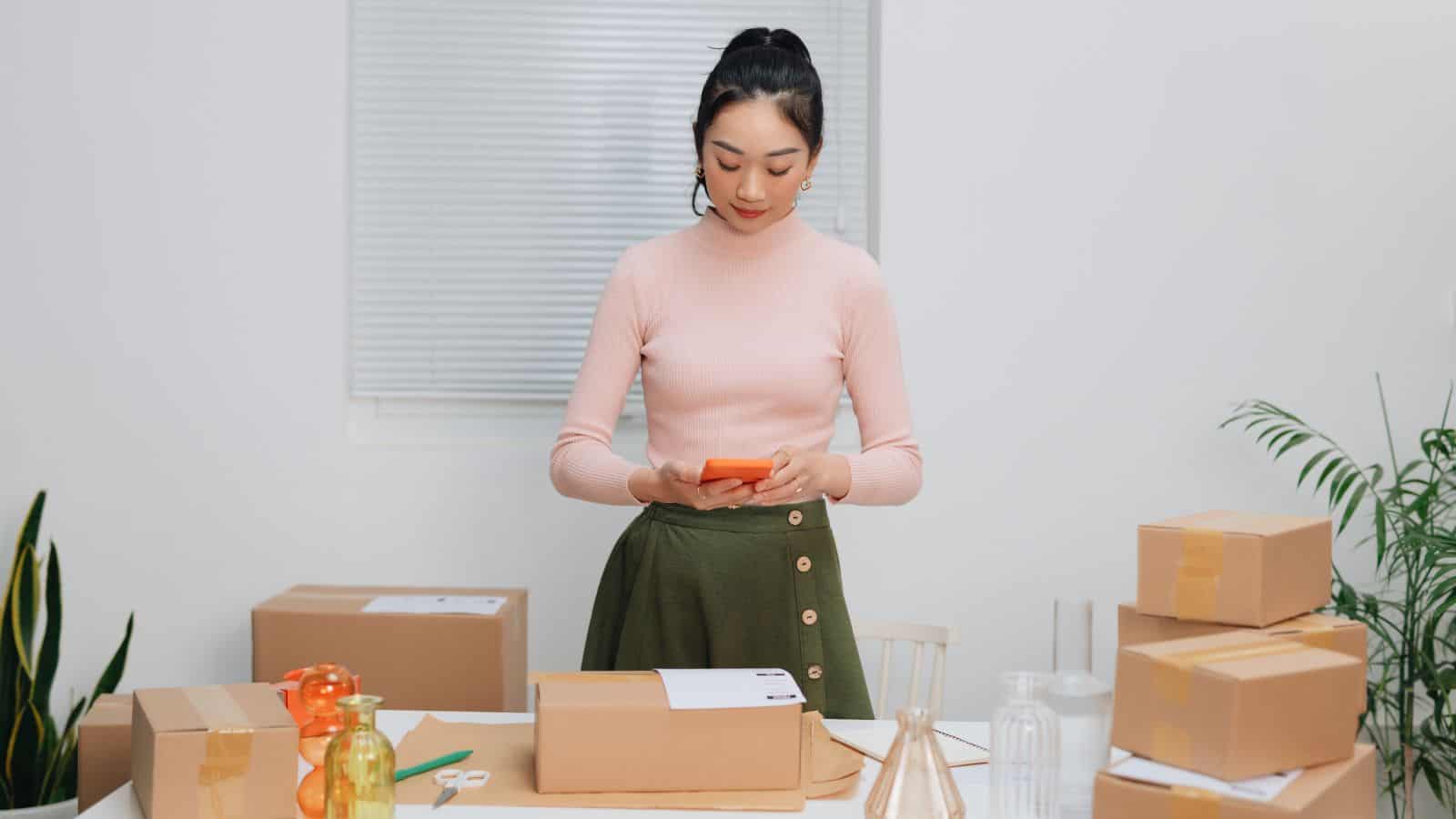 A person in a pink sweater and green skirt stands at a table, using a smartphone. The table is surrounded by packages and shipping materials, with plants visible in the background.