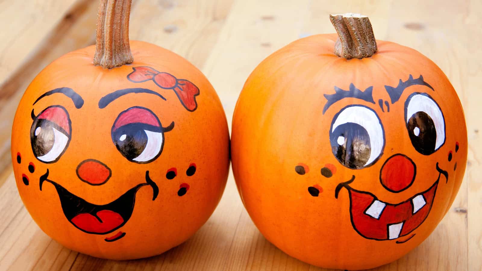 Two pumpkins with painted cartoon faces sit on a wooden surface. The left pumpkin has large eyes, arched eyebrows, and a bow, while the right one features big eyes, bushy eyebrows, and prominent teeth. Both have red cheeks and smiles.