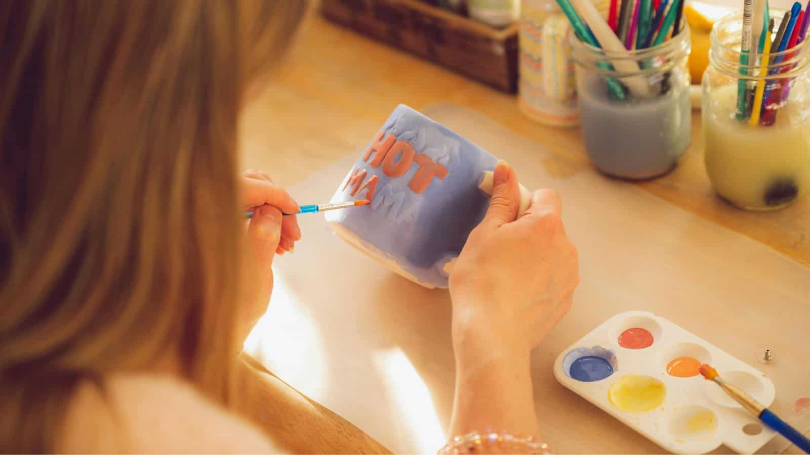 A person is painting a ceramic mug with the phrase "HOT MAMA" using a small brush. The paint palette has four colors: white, orange, yellow, and blue. Various art supplies, including brushes, are organized in jars on the desk.