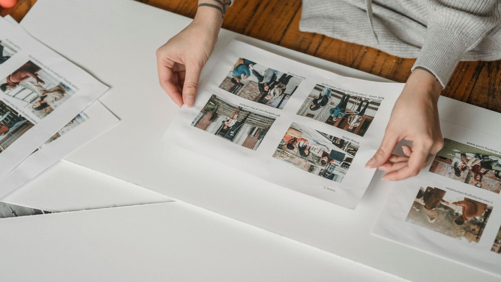 A person is handling printed photos on a table. The photos show various images with people. The person wears a gray sweater, and there are several sheets of paper around. The tabletop is wooden.