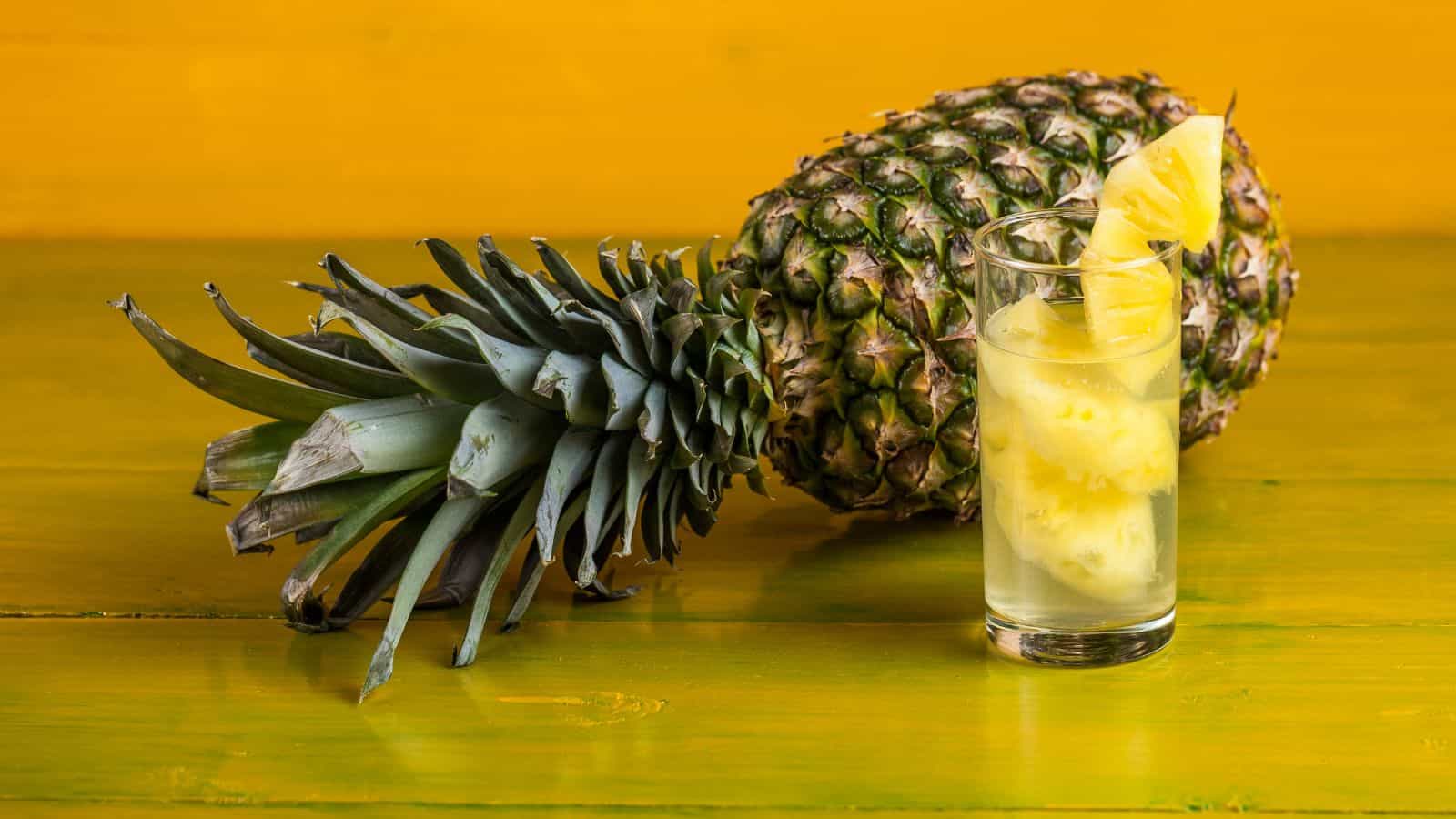 A whole pineapple lies on a wooden surface with a yellow background. In front of it, a clear glass filled with pineapple juice and slices of pineapple is placed.