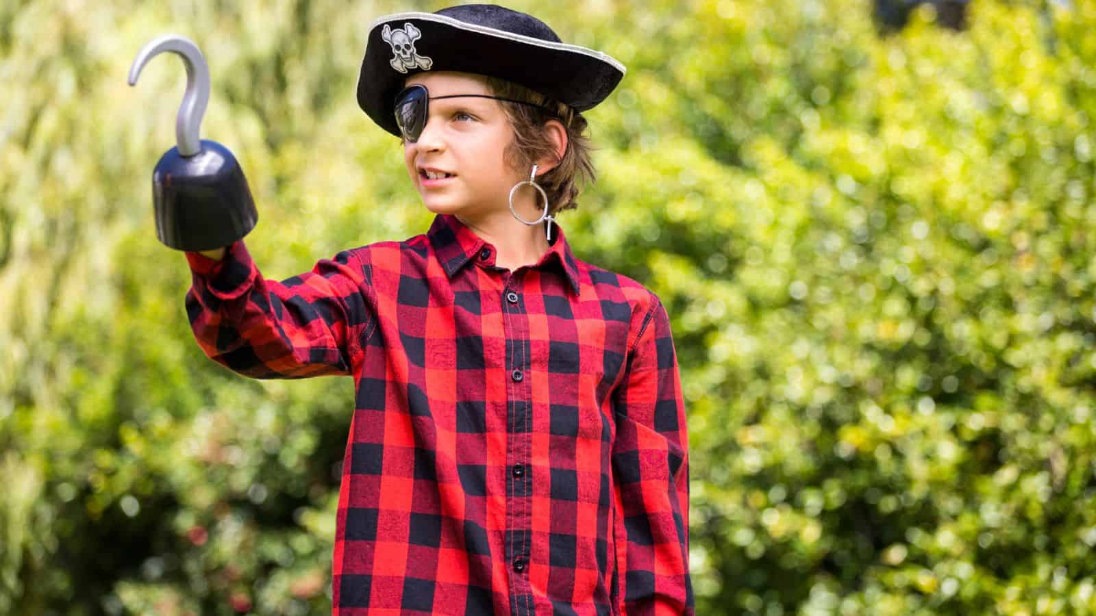 A child wearing a pirate costume, including a hat with a skull emblem, an eye patch, and a hook, stands outdoors. They are dressed in a red and black checkered shirt, with greenery in the background.