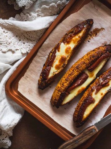 Baked plantains with cheese on a parchment-lined baking tray. A knife rests on the tray, and a white fabric with lace detailing is partially visible beside it.