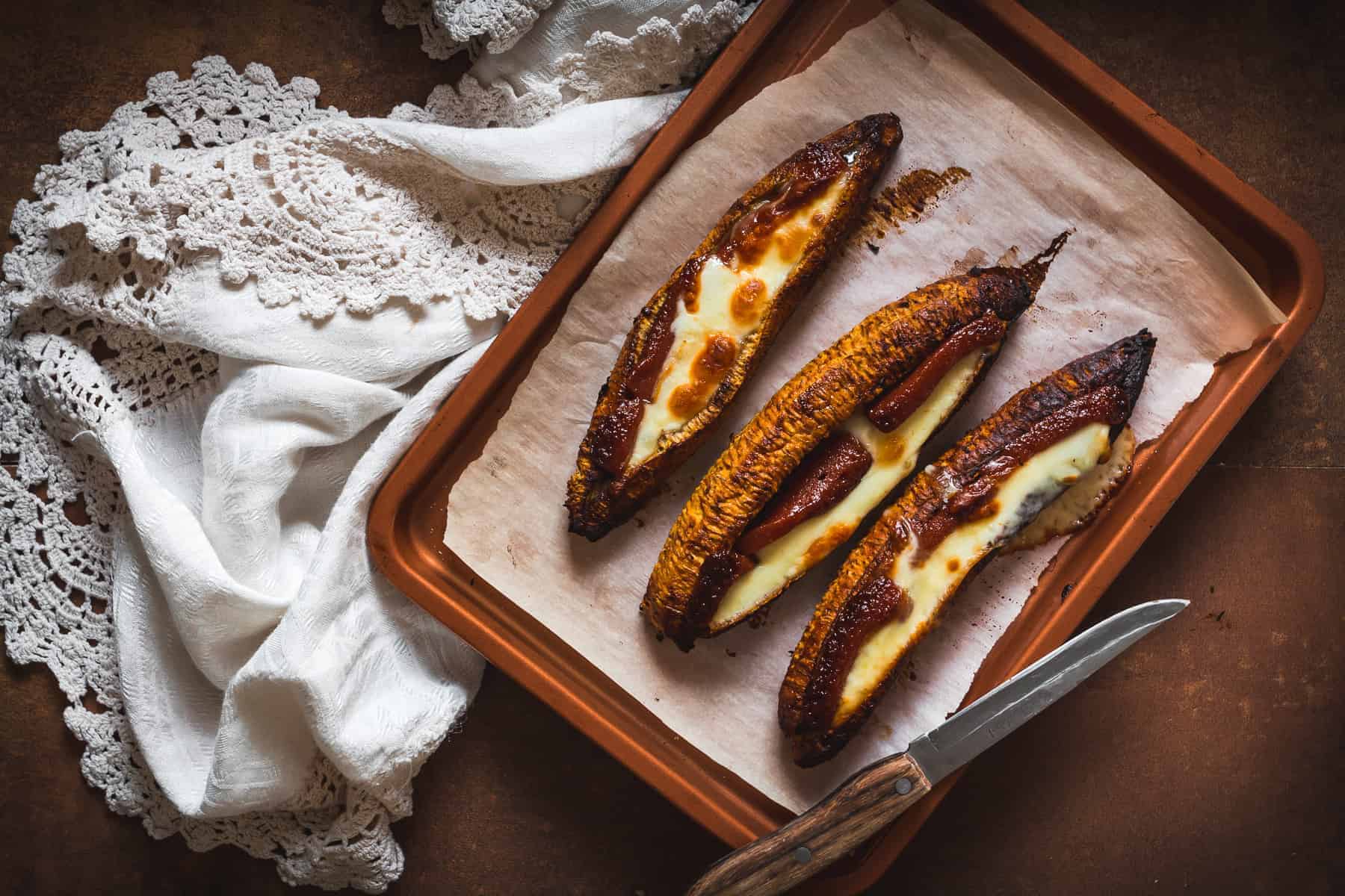 Baked plantains with cheese on a parchment-lined baking tray. A knife rests on the tray, and a white fabric with lace detailing is partially visible beside it.