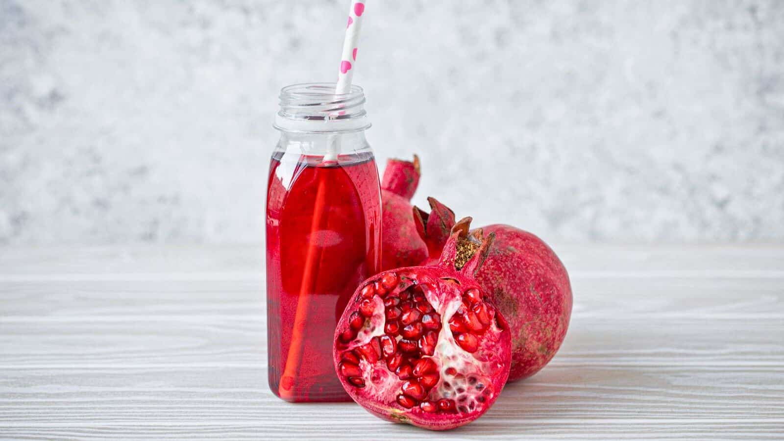 A glass bottle filled with pomegranate juice and a pink polka dot straw stands next to a whole pomegranate and a halved one, showing the red seeds inside, on a light wooden surface with a gray speckled background.