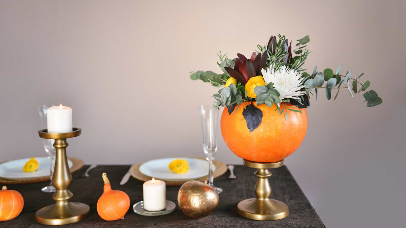 A table setting with a fall theme, featuring a pumpkin vase centerpiece filled with greenery and flowers. Surrounding the arrangement are lit candles on gold stands, small pumpkins, a gold pear, and white plates with yellow accents on a dark tablecloth.