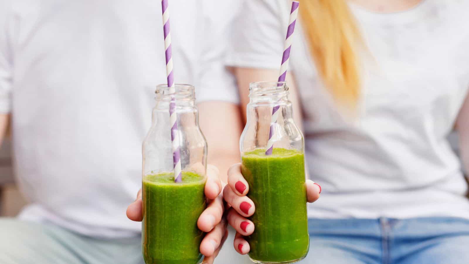 Two people holding glass bottles filled with green smoothies. The bottles have striped purple and white straws, and both individuals are wearing white shirts. One person has painted nails. The background is out of focus.