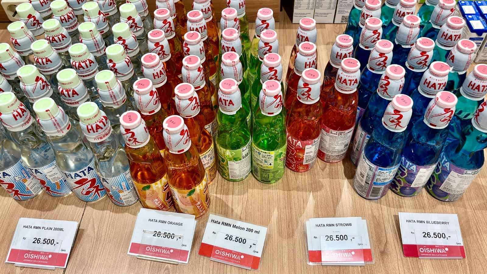 A display of various flavored Hatakosen Ramune drinks on a wooden surface. Bottles are arranged in rows by flavor: plain, orange, melon, lemon, and blueberry. Price tags below each row indicate they cost 26,500 units each.