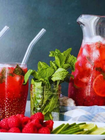A pitcher and two glasses of red fruit punch with ice and clear straws sit on a table. A jar of fresh mint is beside the drinks. A plate of raspberries, cucumber slices, and lime wedges is in the foreground, with a cloth draped under the pitcher.