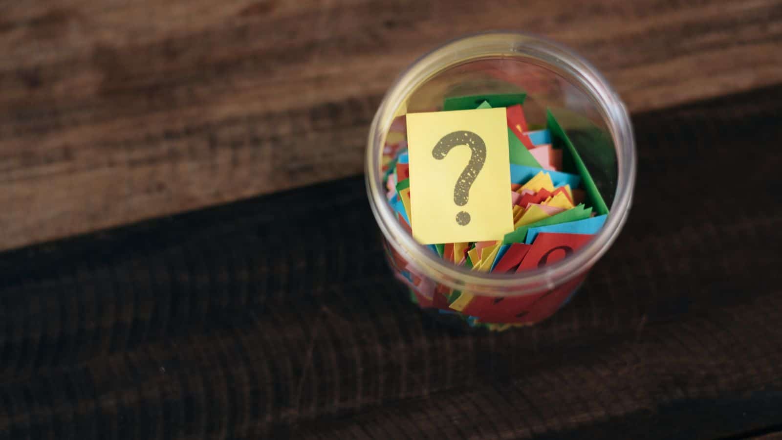 A clear jar filled with colorful paper slips on a wooden surface. A yellow slip with a large question mark is placed on top.