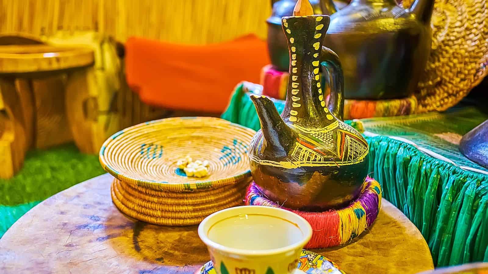 A traditional Ethiopian coffee setup is displayed, featuring a painted coffee pot and a small cup on a wooden table. A woven basket holds white decorative beads, and vibrant textiles adorn the background.