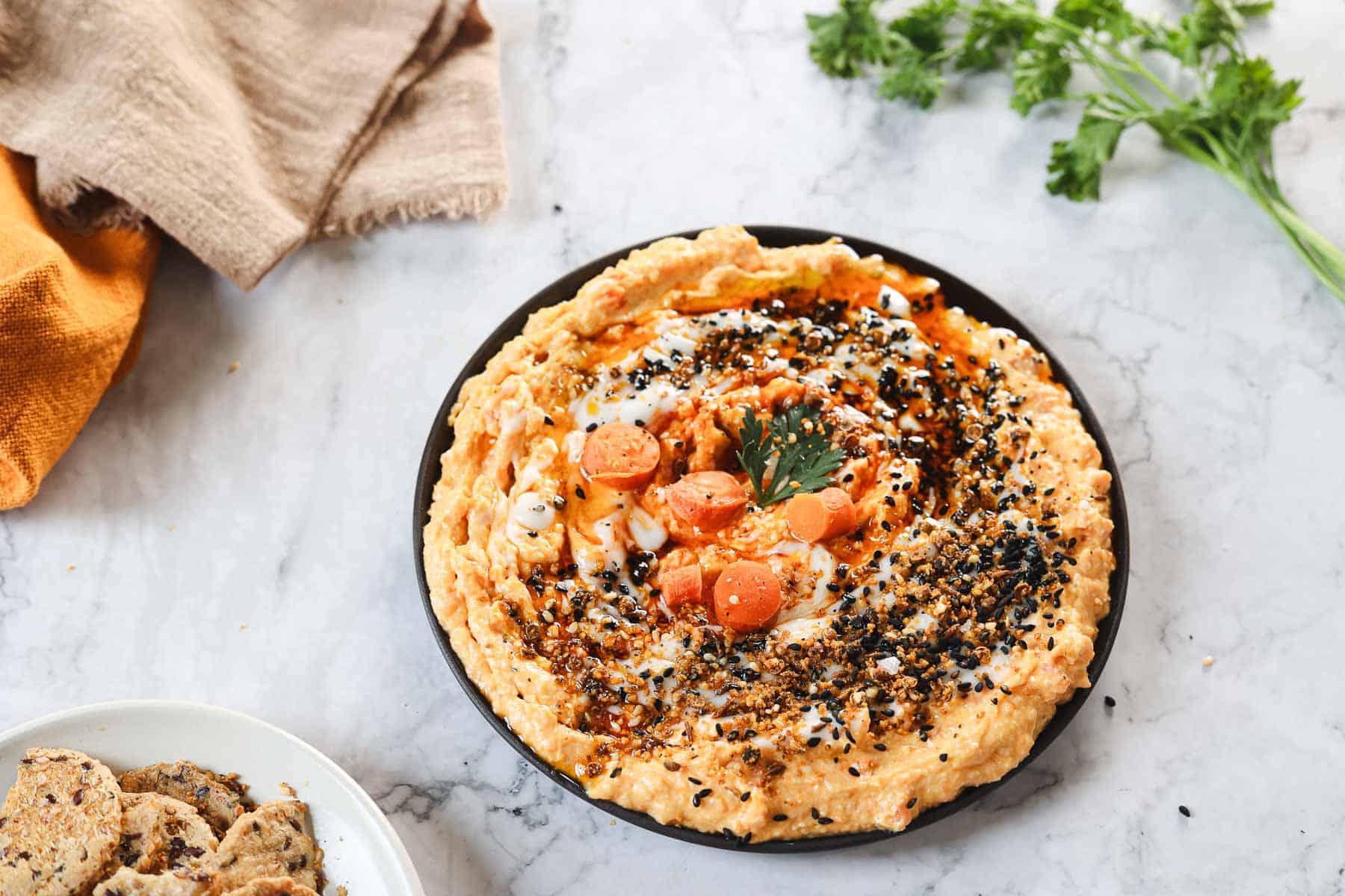 A plate of creamy carrot dip, stylishly garnished with sliced carrots, a parsley leaf, black sesame seeds, and spices. It rests on a marble surface alongside a beige cloth and fresh parsley sprigs. A bowl brimming with round crackers is partially visible nearby.