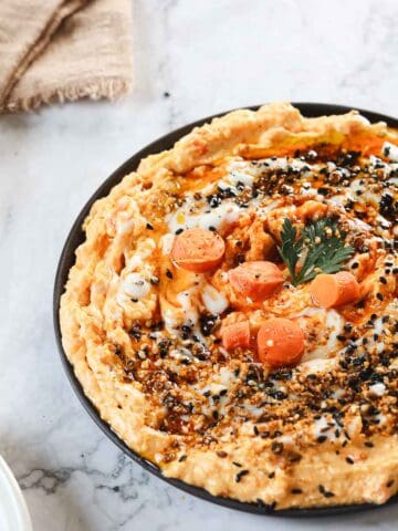 A plate of carrot dip hummus topped with roasted carrots, black sesame seeds, dukkah, and a drizzle of sauce sits on a marble countertop. A sprig of parsley garnishes the center. A cloth napkin and a small plate of crackers are nearby.