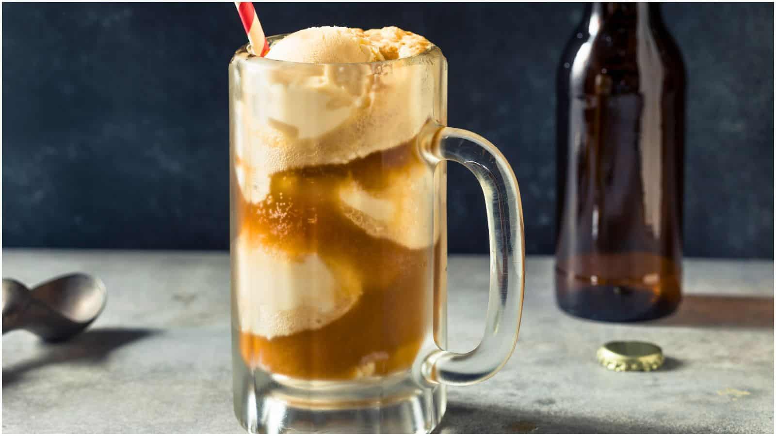 A glass mug filled with root beer float, featuring scoops of vanilla ice cream and a red-striped straw. A brown bottle stands in the background on a gray surface, accompanied by a bottle cap and an ice cream scoop.