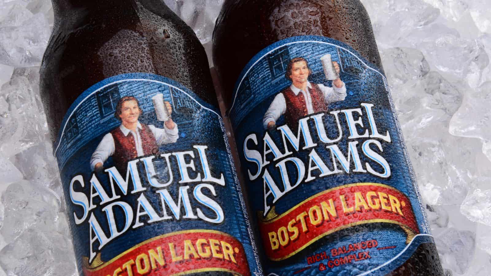 Two bottles of Samuel Adams Boston Lager are placed on ice. The labels feature a historical figure holding a beer mug and the text "Rich, Balanced & Complex." The bottles are slightly angled, with condensation visible on their surfaces.