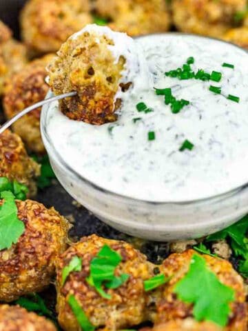 A skillet filled with cooked meatballs garnished with parsley surrounds a small bowl of creamy white sauce topped with chopped chives. A wooden-handled fork holds a meatball dipped in the sauce.