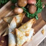 Four golden-brown savoury pastries shaped as phyllo triangles rest on a wooden board, surrounded by mushrooms, parsley, and a shallot.